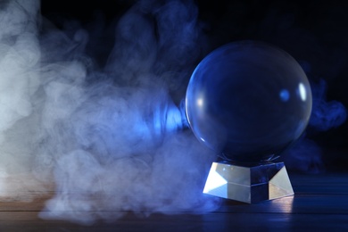 Photo of Magic crystal ball on wooden table and smoke against dark background, space for text. Making predictions