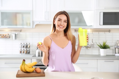 Photo of Young woman holding bottle of protein shake near table with ingredients in kitchen