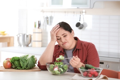 Unhappy woman eating vegetable salad at table in kitchen. Healthy diet