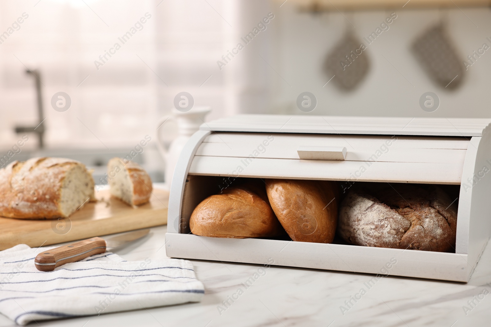 Photo of Wooden bread basket with freshly baked loaves and knife on white marble table in kitchen