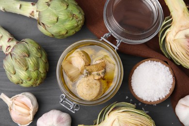 Jar of delicious artichokes pickled in olive oil and ingredients on dark grey wooden table, flat lay