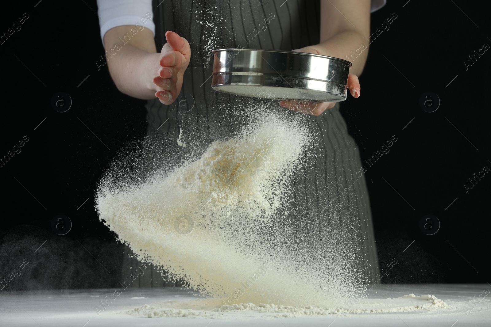 Photo of Woman sieving flour at table against black background, closeup