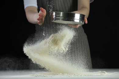 Photo of Woman sieving flour at table against black background, closeup