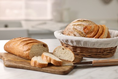 Photo of Wicker bread basket with freshly baked loaves and knife on white marble table in kitchen