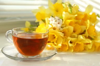 Photo of Cup of aromatic tea and beautiful yellow daffodils on windowsill