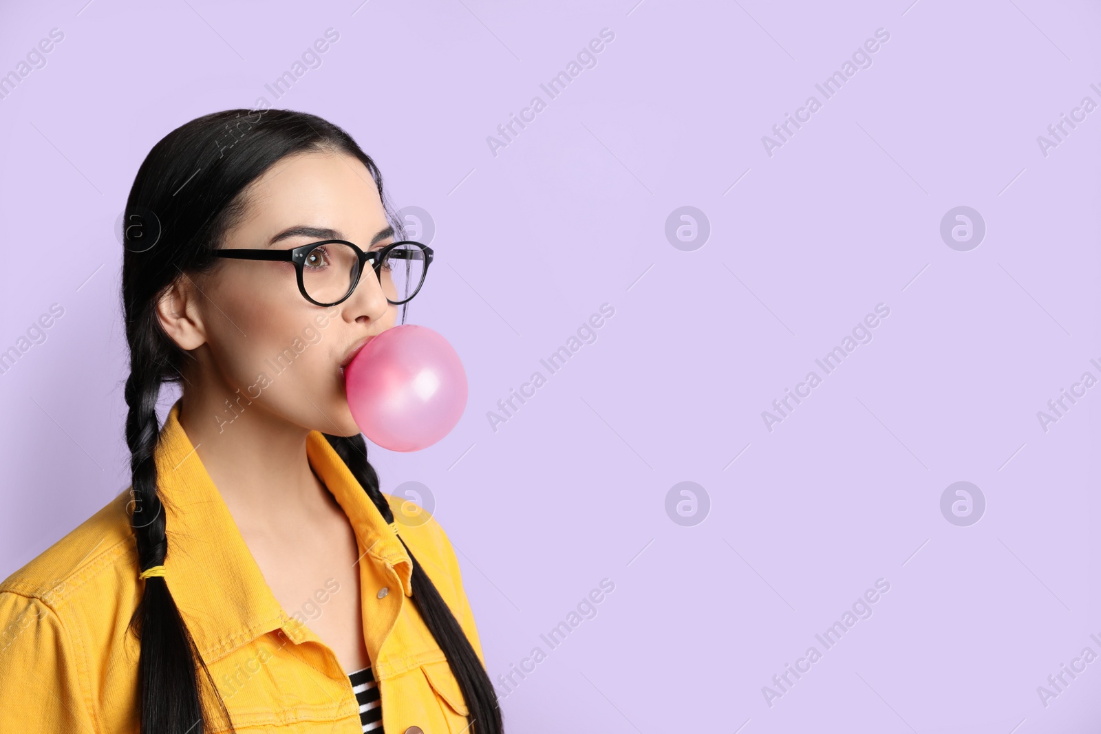 Photo of Fashionable young woman with braids blowing bubblegum on lilac background, space for text