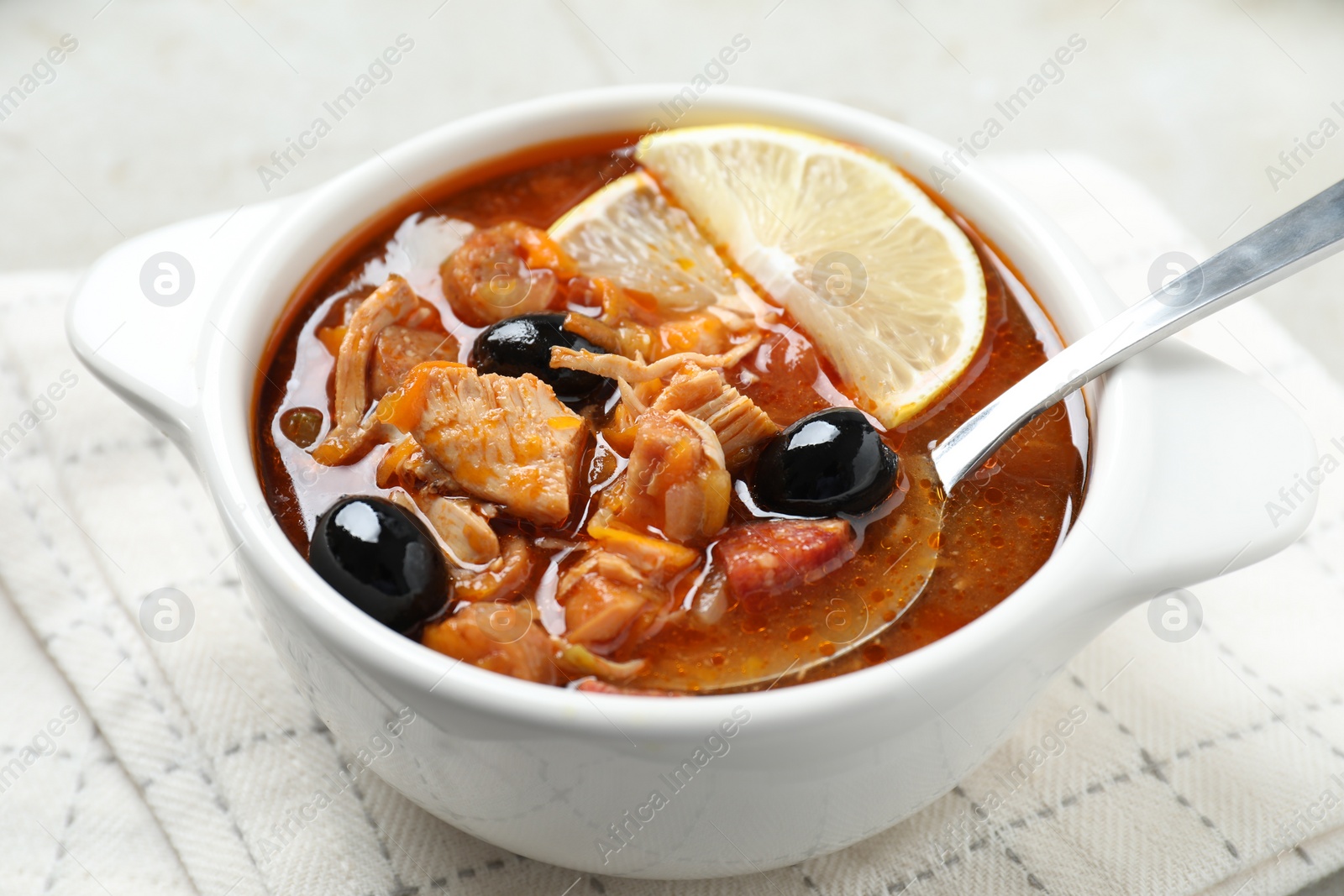 Photo of Meat solyanka soup with sausages, olives, vegetables and spoon in bowl on white table, closeup