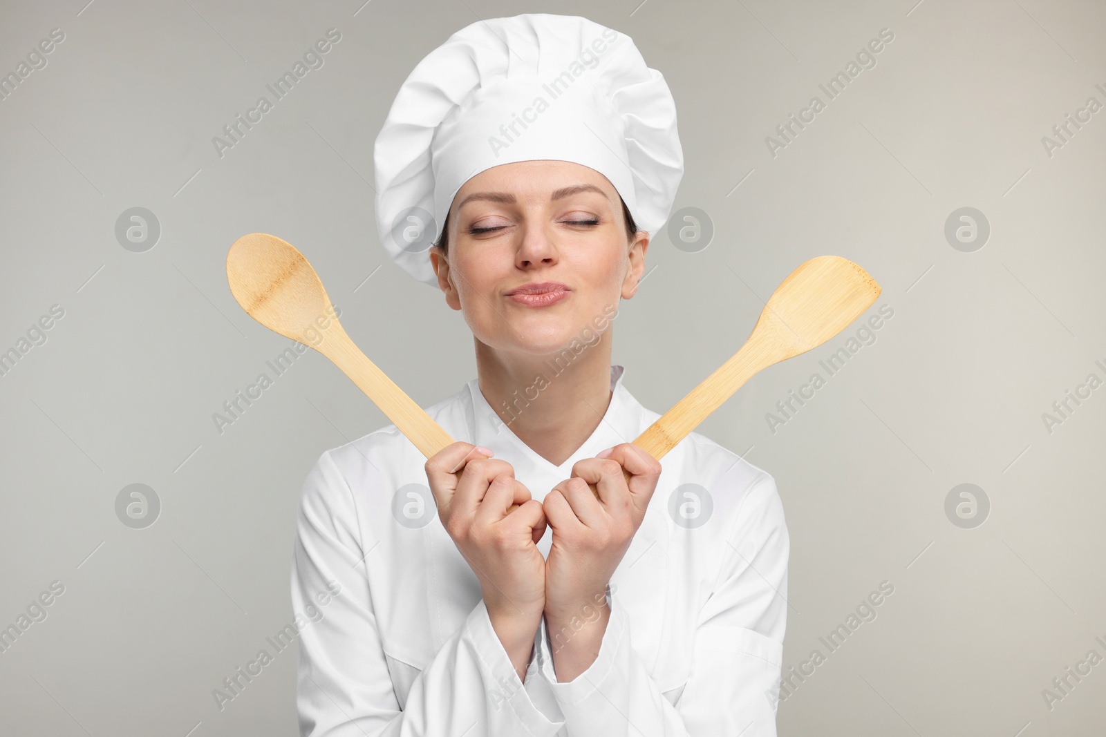 Photo of Happy woman chef in uniform holding wooden spoon and spatula on grey background