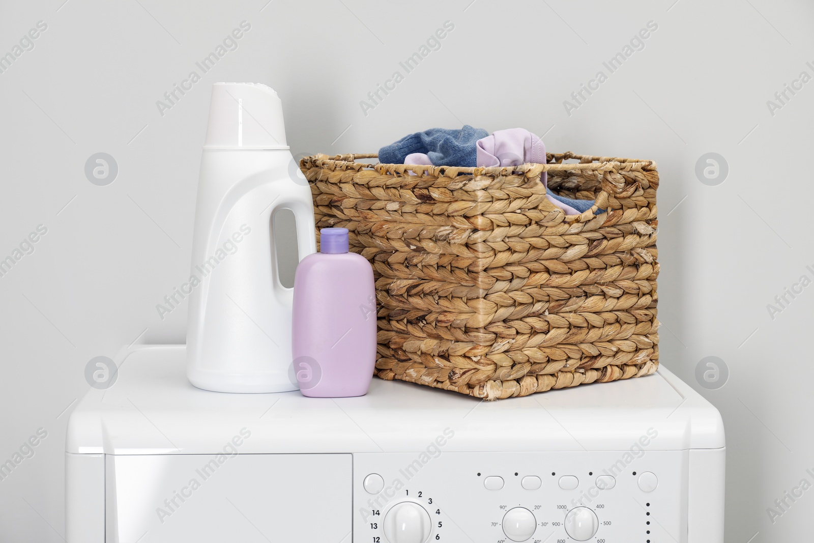 Photo of Baby clothes in wicker basket and laundry detergents on washing machine near light wall