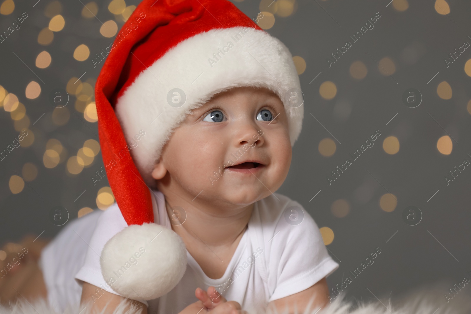 Photo of Cute baby in Santa hat on fluffy carpet against blurred lights. Christmas celebration