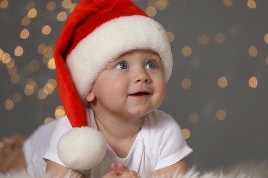 Cute baby in Santa hat on fluffy carpet against blurred lights. Christmas celebration