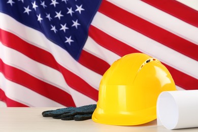 Photo of Yellow hard hat, gloves and paper on white wooden table against American flag, space for text