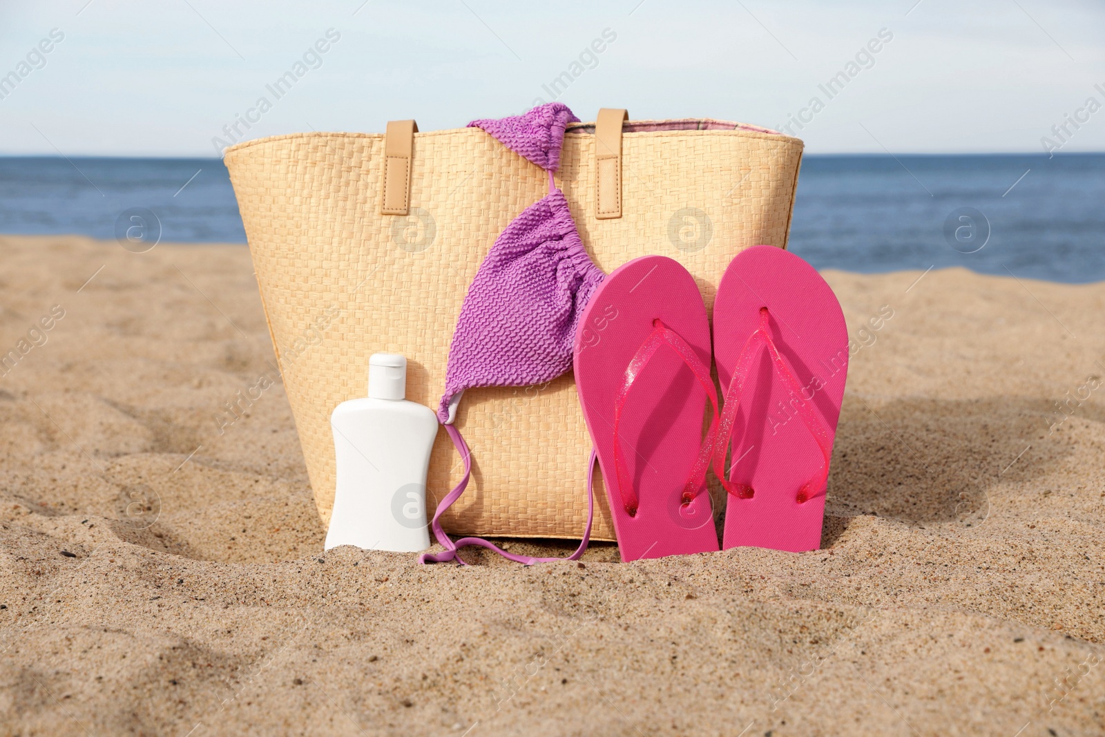 Photo of Summer bag with slippers, sunscreen and bikini top on sand near sea
