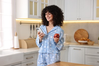 Beautiful young woman in stylish pyjama with smartphone and fresh apple in kitchen