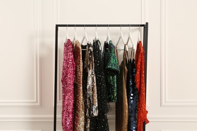 Photo of Clothing rack with colorful sequin party dresses on hangers near white wall indoors