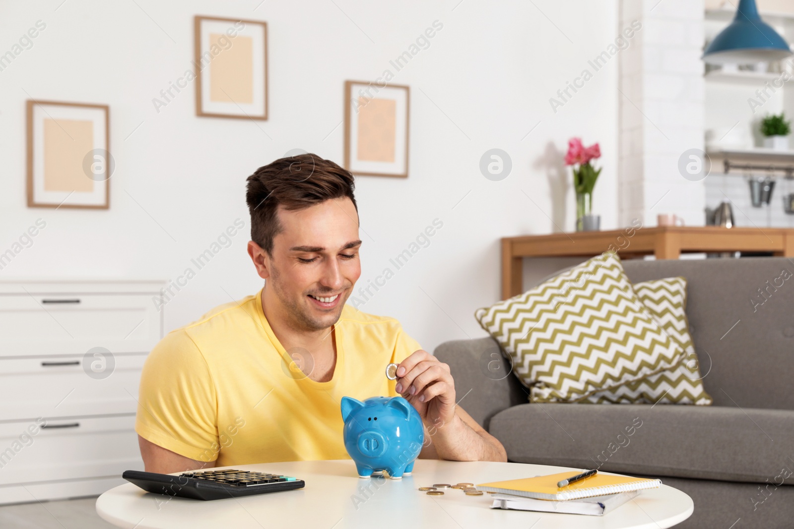 Photo of Happy man putting coin into piggy bank at table in living room. Saving money