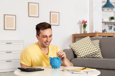 Happy man putting coin into piggy bank at table in living room. Saving money