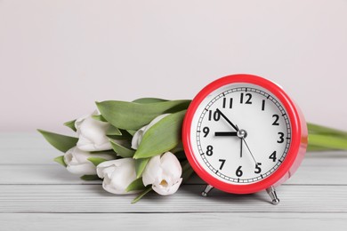 Photo of Red alarm clock and beautiful tulips on white wooden table against light background. Spring time