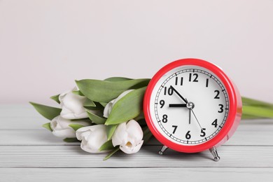 Red alarm clock and beautiful tulips on white wooden table against light background. Spring time