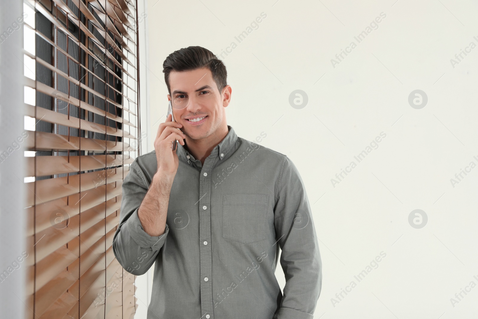 Photo of Handsome man talking on mobile phone at window indoors. Space for text