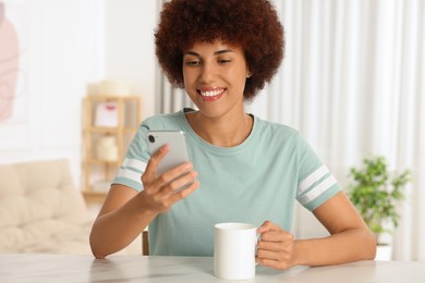 Happy young woman with cup of drink using smartphone at table indoors