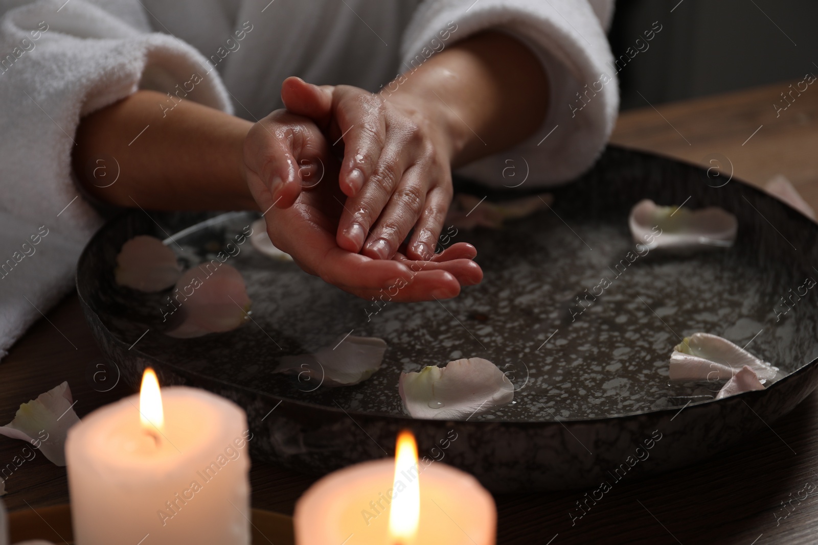 Photo of Woman soaking her hands in bowl of water and flower petals at table, closeup. Spa treatment