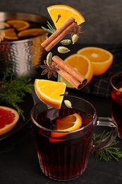 Cut orange and different spices falling into glass cup of mulled wine on table