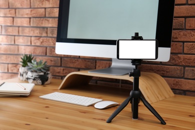 Tripod with smartphone near computer on wooden table indoors. Mockup for design