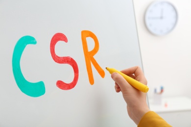 Woman writing abbreviation CSR on magnetic whiteboard, closeup. Corporate social responsibility