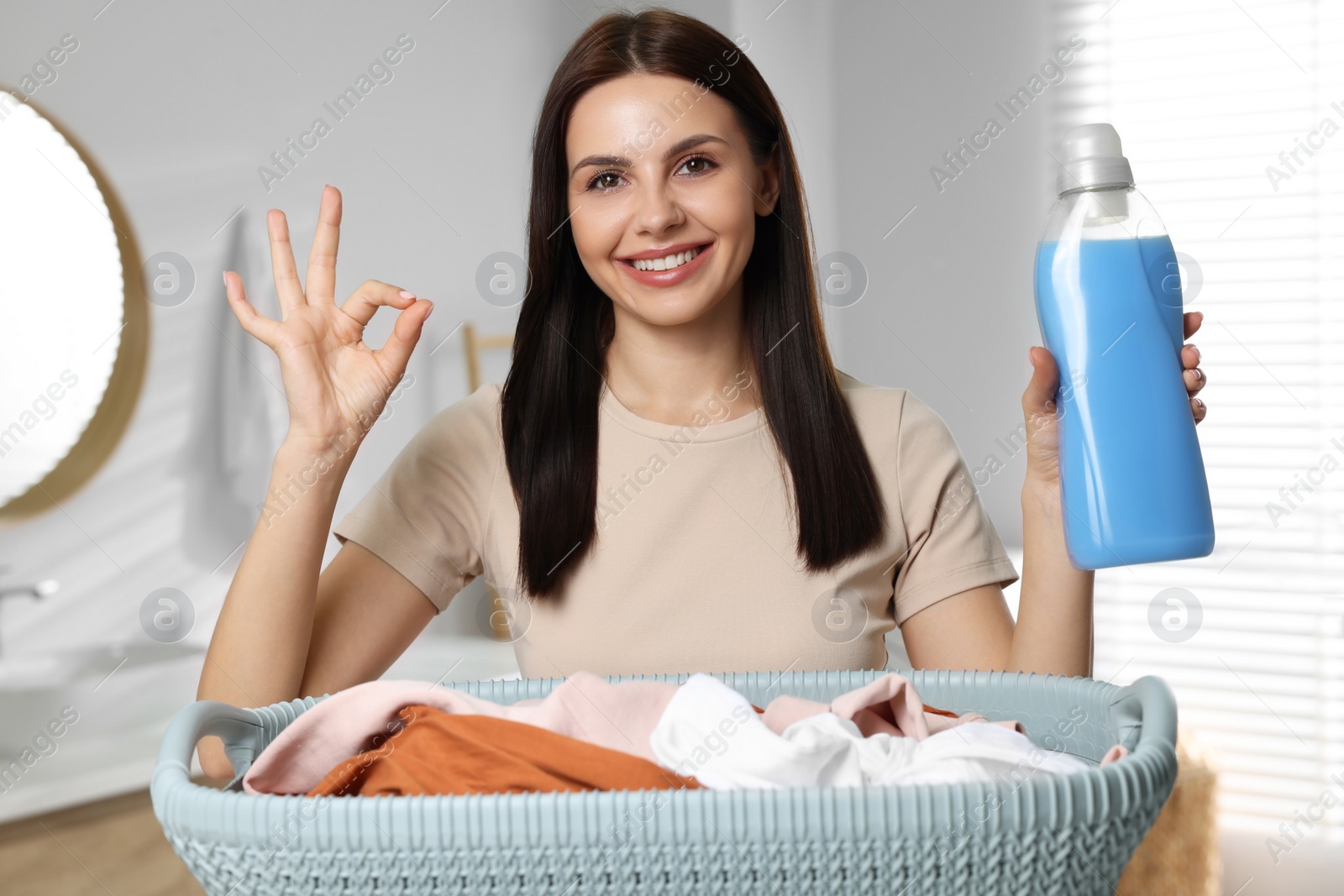 Photo of Woman holding fabric softener near basket with dirty clothes and showing OK gesture in bathroom