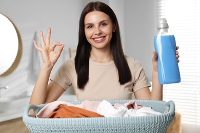 Photo of Woman holding fabric softener near basket with dirty clothes and showing OK gesture in bathroom