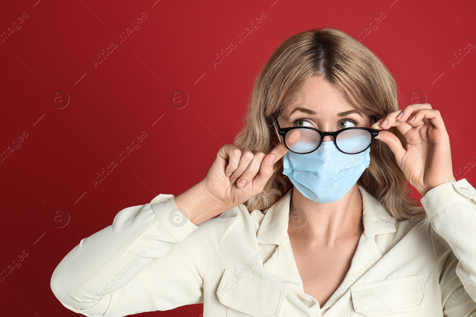 Photo of Woman wiping foggy glasses caused by wearing medical mask on red background