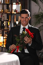 Photo of Happy man with roses waiting for his girlfriend in restaurant on Valentine's day