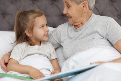 Photo of Cute girl and her grandmother reading book on bed at home