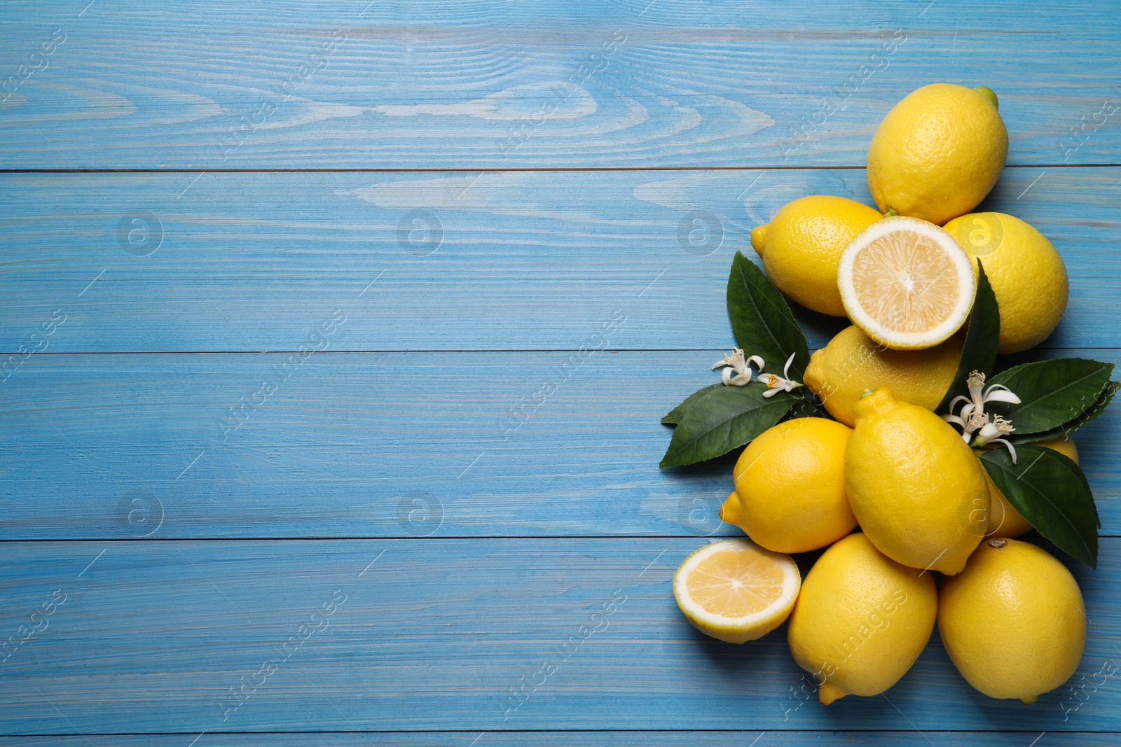 Photo of Many fresh ripe lemons with green leaves and flowers on blue wooden table, flat lay. Space for text