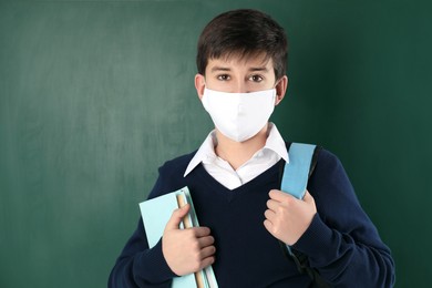Boy wearing protective mask with backpack and books near chalkboard. Child safety