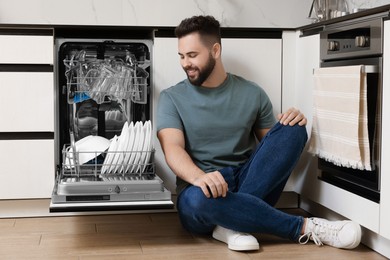 Photo of Smiling man sitting near open dishwasher in kitchen