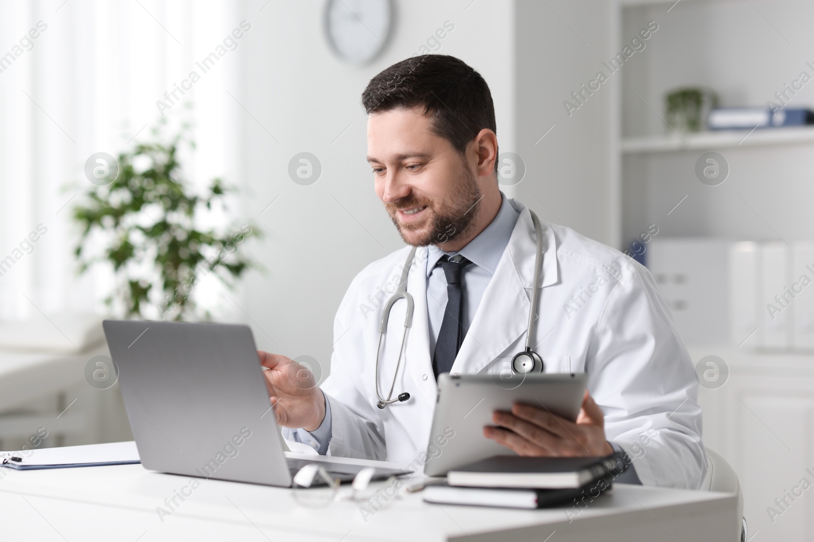 Photo of Smiling doctor with gadgets having online consultation at table in clinic