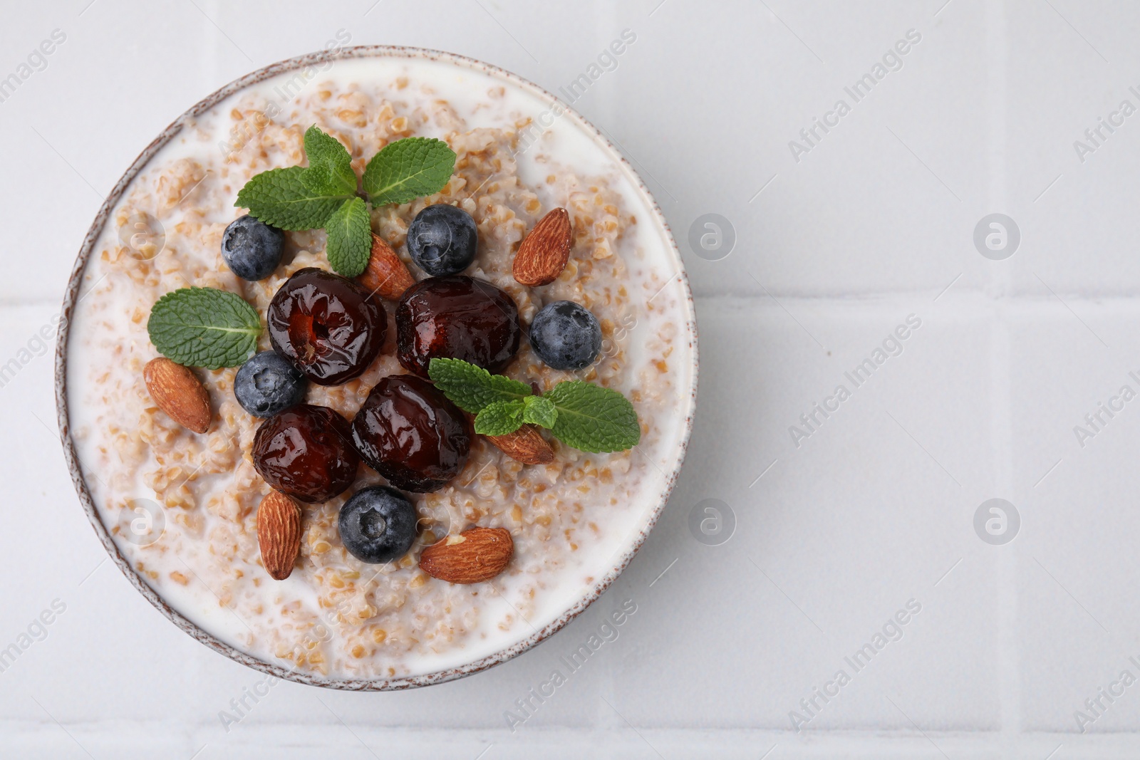 Photo of Tasty wheat porridge with milk, dates, blueberries and almonds in bowl on white table, top view. Space for text