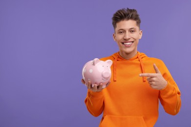 Photo of Happy man pointing at piggy bank on purple background. Space for text