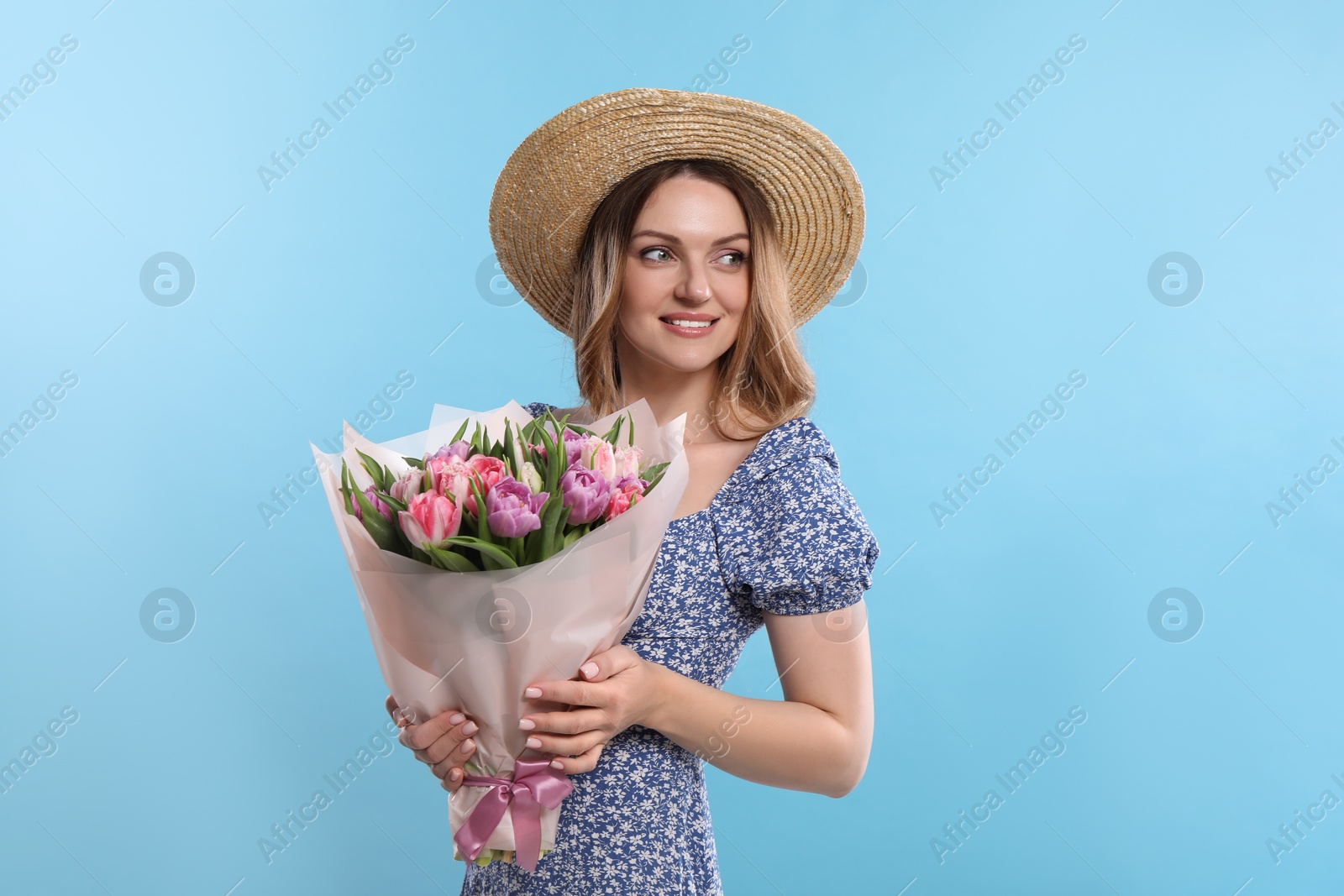Photo of Happy young woman in straw hat holding bouquet of beautiful tulips on light blue background