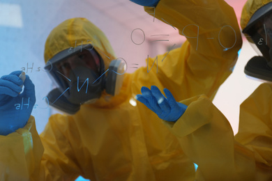 Scientists in chemical protective suits writing formula on glass board at laboratory. Virus research