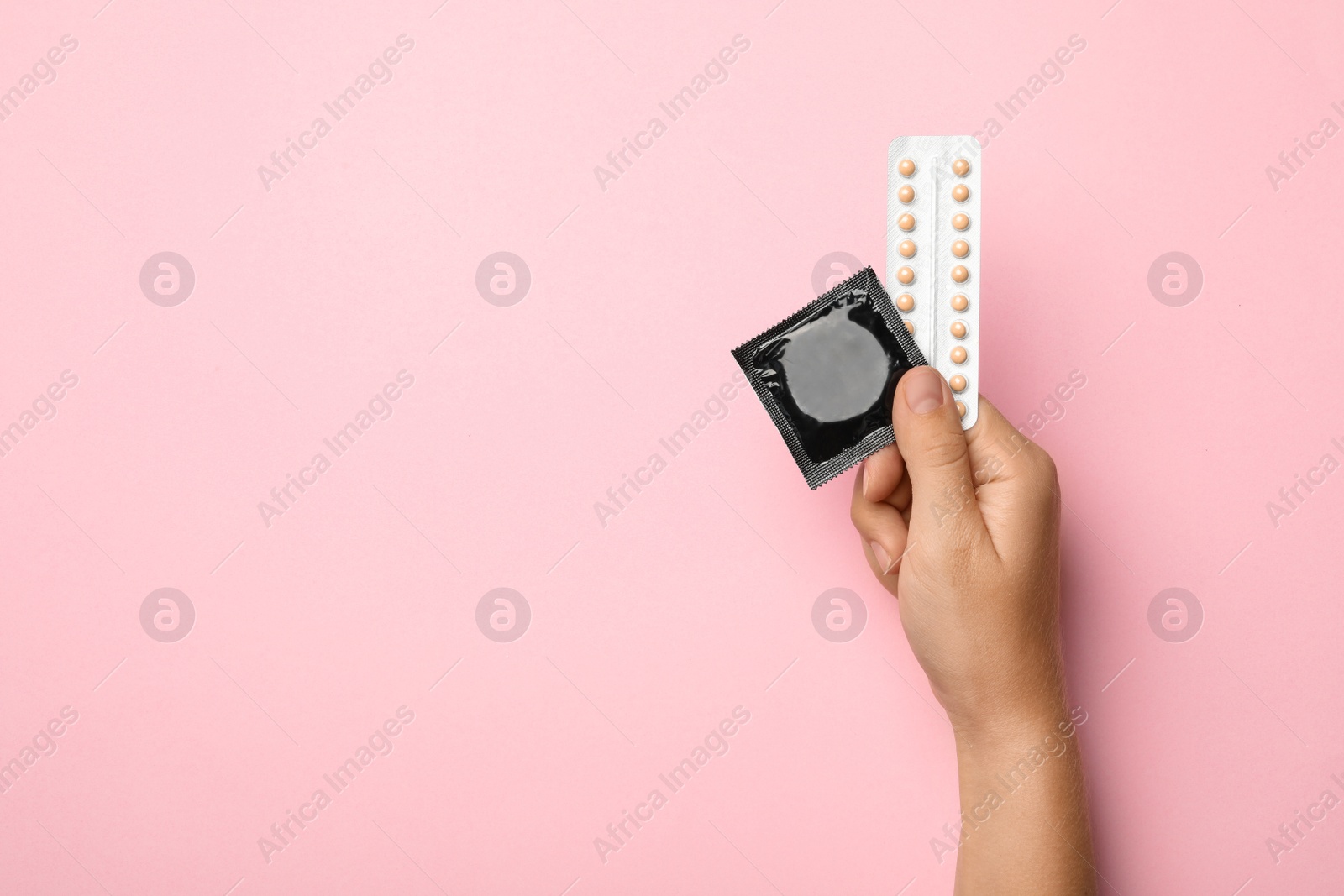 Photo of Woman holding condom and birth control pills on pink background, top view with space for text. Safe sex