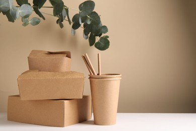 Photo of Eco friendly food packaging. Paper containers, straws and eucalyptus branches on white table against beige background. Space for text