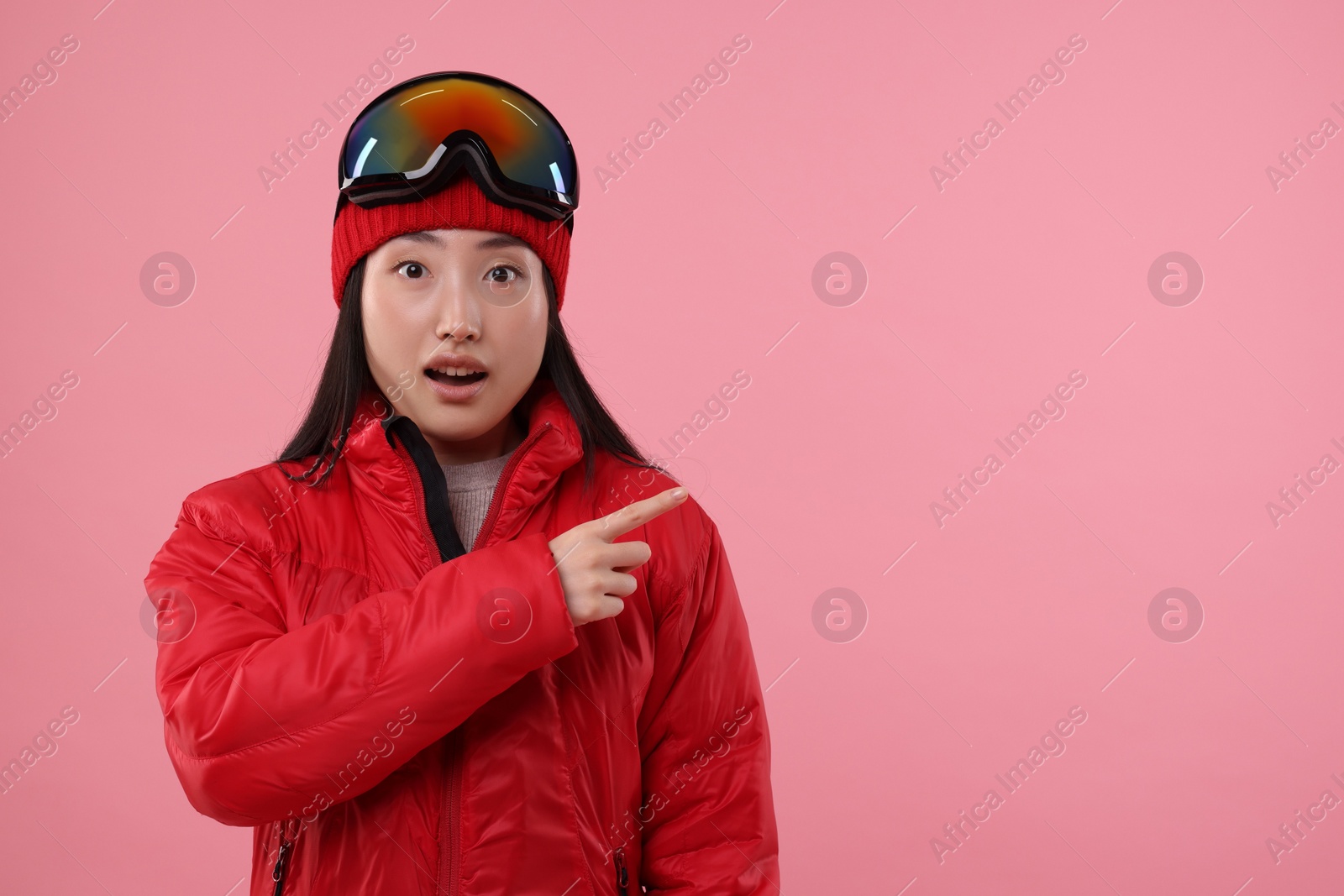 Photo of Surprised woman in winter sportswear pointing at something on pink background. Space for text