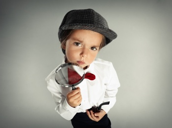 Photo of Cute little child in hat with smoking pipe and magnifying glass playing detective on grey background