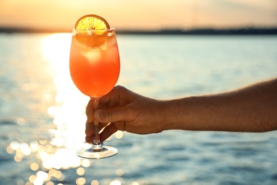 Photo of Man with glass of fresh summer cocktail near water at sunset, closeup