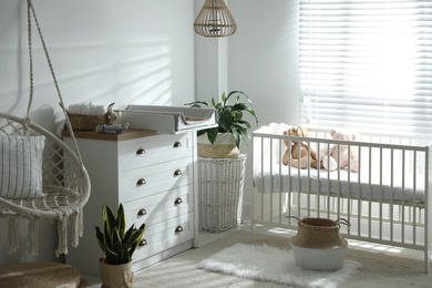 Photo of Chest of drawers with changing tray and pad near comfortable cradle in baby room. Interior design