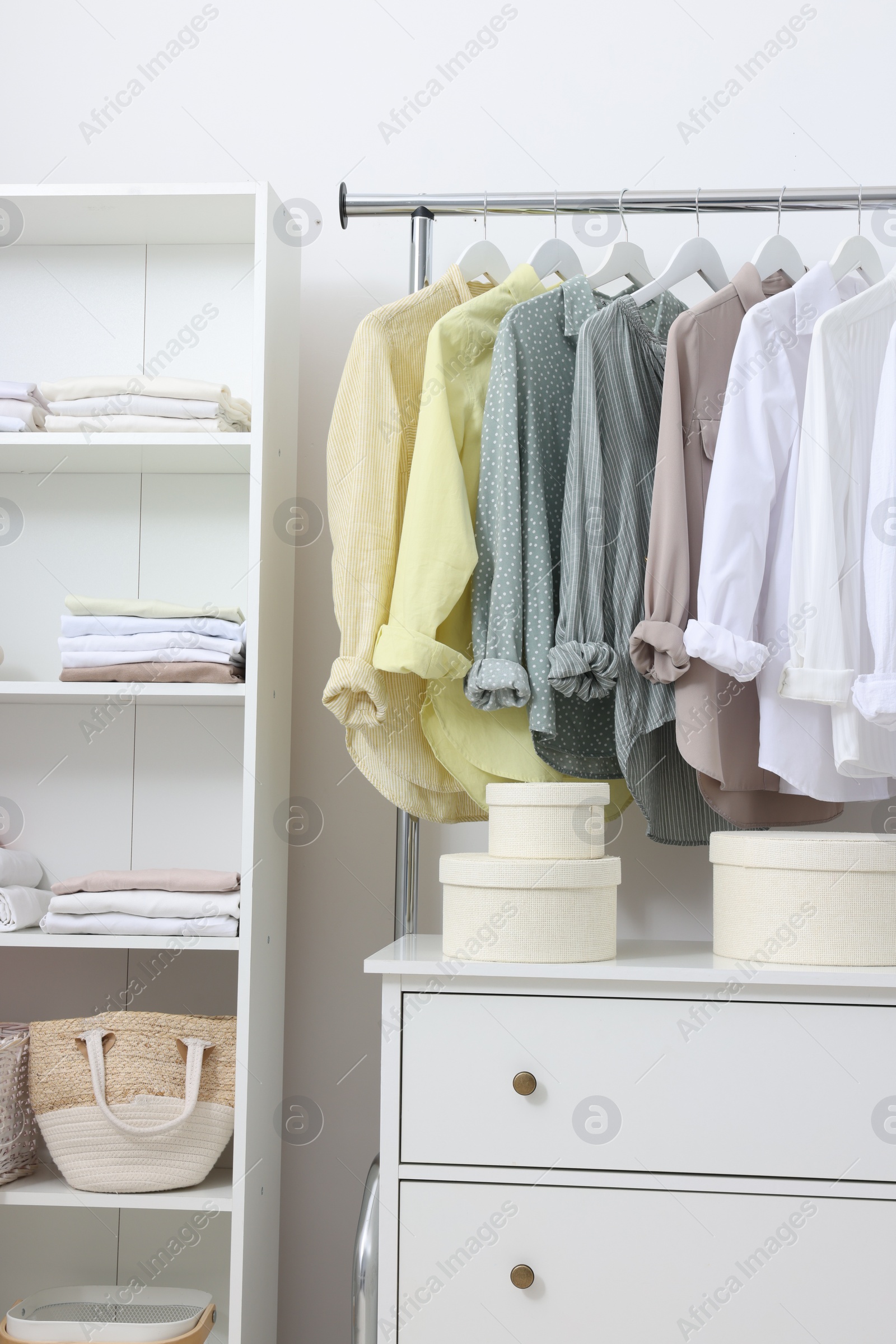 Photo of Wardrobe organization. Rack with different stylish clothes, shelving unit and chest of drawers near white wall