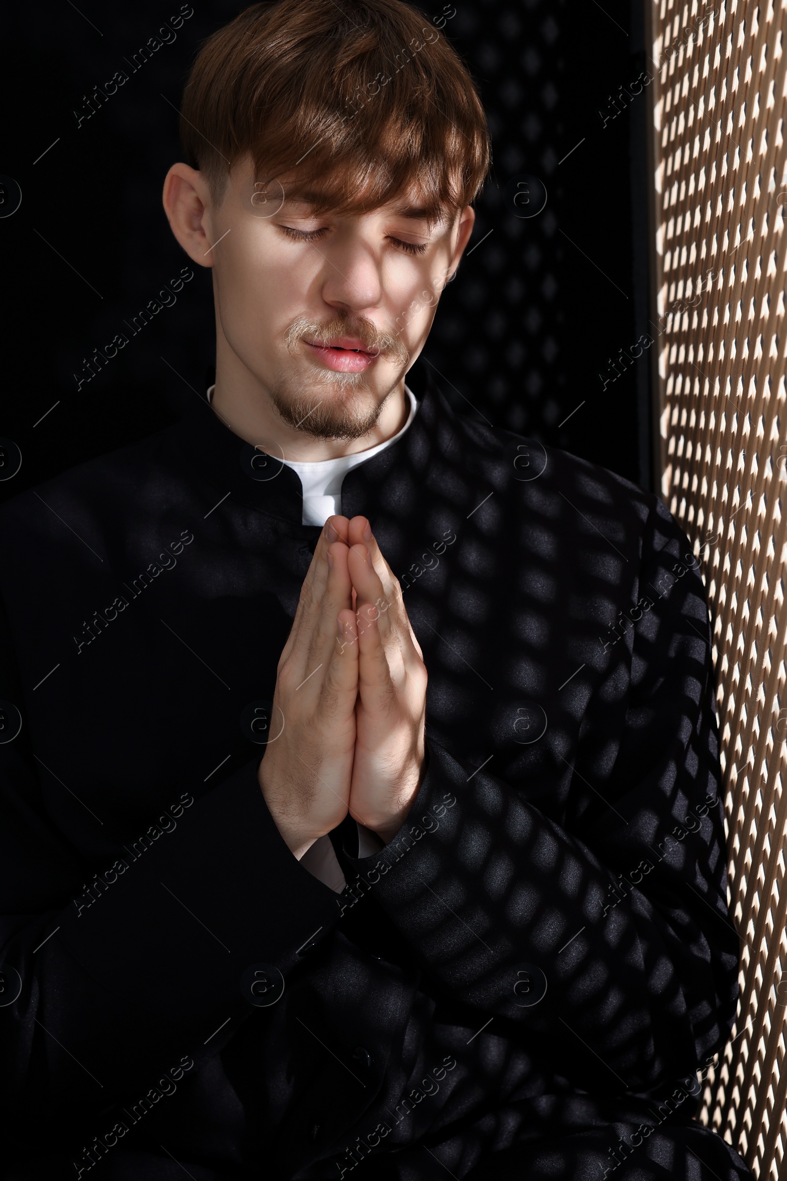 Photo of Catholic priest praying near wooden window in confessional booth